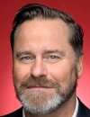 A head shot of Senator Peter Whish Wilson wearing a dark suit and white shirt. The background is red.