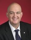 An image of Senator Slade Brockman wearing a dark suit with a red background.
